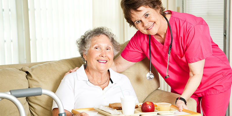 The doctor, dressed in pink, holds a plate of food for the elderly woman sitting on a sofa while both smile and pose for the camera