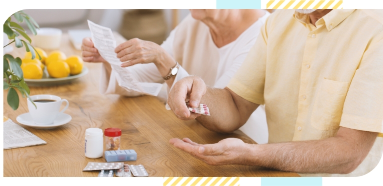 Two elderly people sitting at their wooden table with pills and pill bottles on it, taking their medication