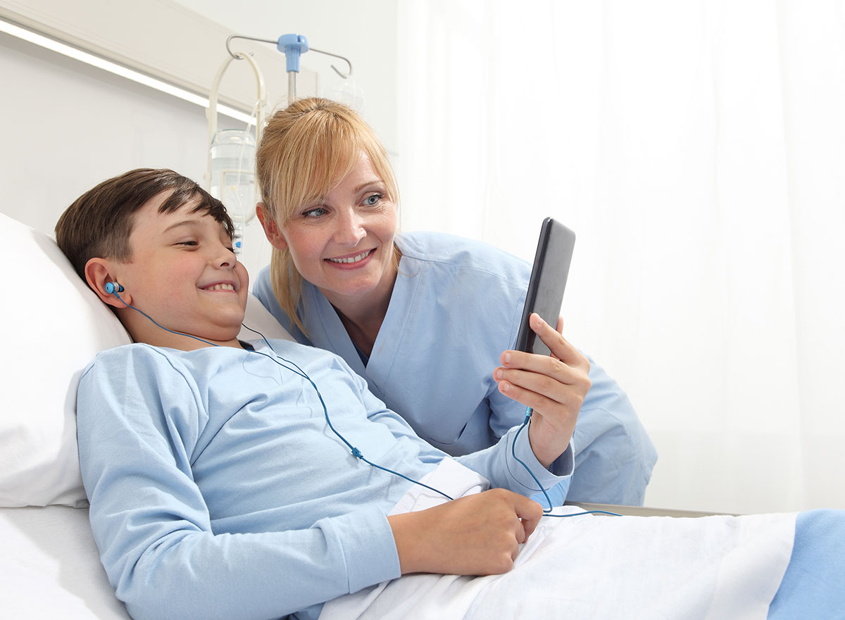 In a hospital environment, a child in a hospital bed holds a mobile phone in his hand while his mother watches the screen of the mobile device.