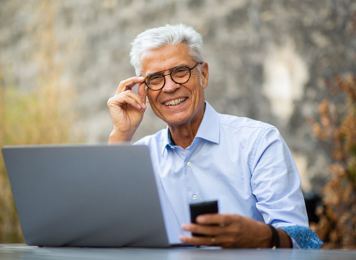Old man smiling and holding his specs as he sits with his laptop and mobile phone