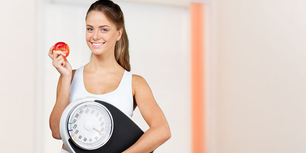 A healthy woman carrying a weighing machine and holding an apple to eat.